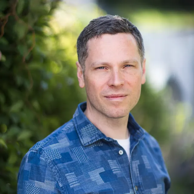 Outdoor portrait of Allan Radcliffe wearing a blue patterned shirt, with green foliage in the background